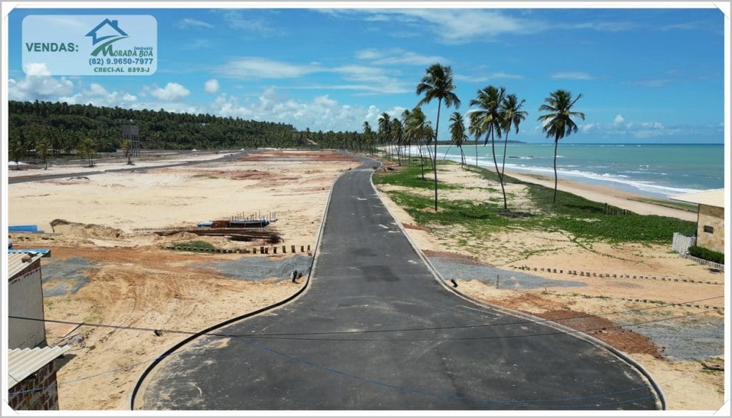 Vista de uma estrada recém-asfaltada que se estende na orla, cercada por palmeiras e áreas de areia, sob um céu azul claro.