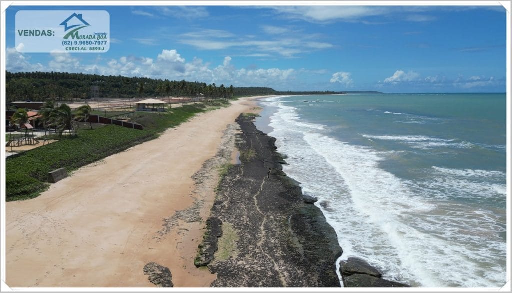 Praia com areia clara, rochas à beira-mar e vegetação à distância, céu azul com nuvens brancas, ambiente tranquilo e natural.