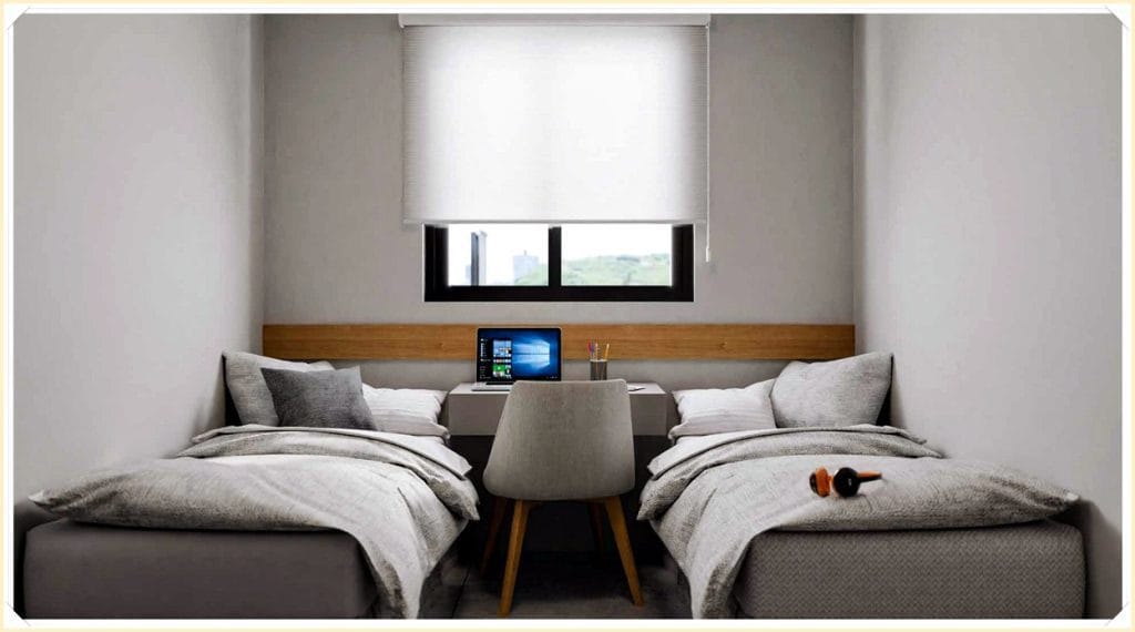 Quarto com duas camas de solteiro, mesa de trabalho com computador, tonalidades neutras e janelas com luz natural.