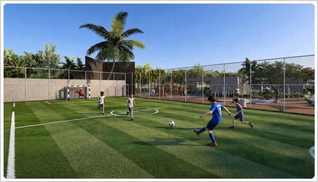Campo de futebol com grama artificial, crianças jogando bola, cercado por plantas e áreas de lazer ao fundo, sob um céu claro.