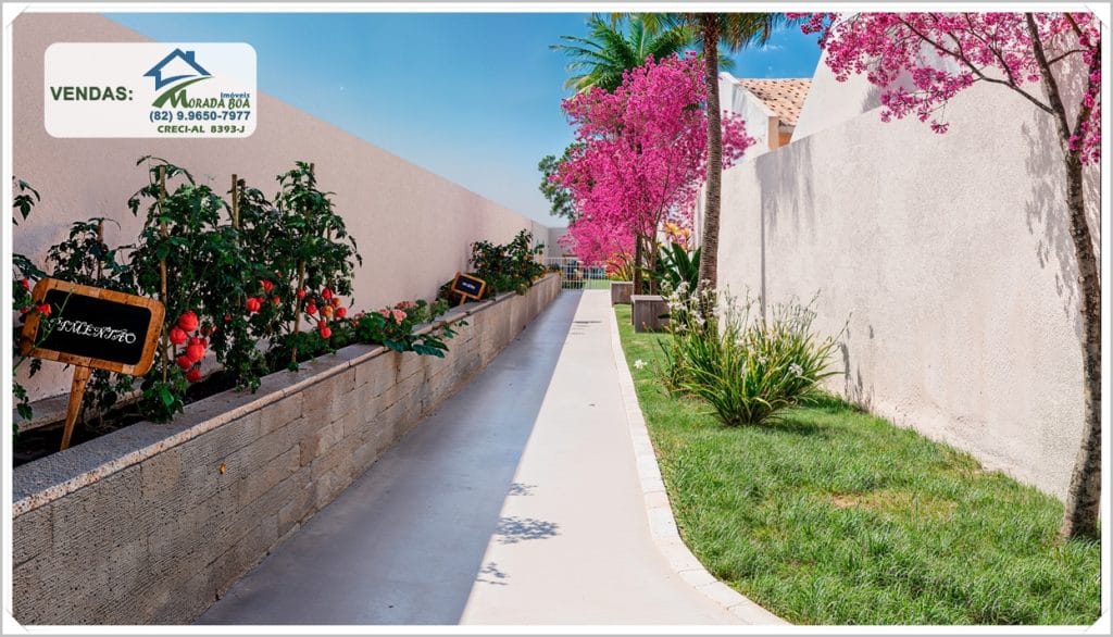 Caminho ajardinado com paredes claras, árvores floridas em rosa e vegetação ao longo, criando um ambiente agradável e convidativo.